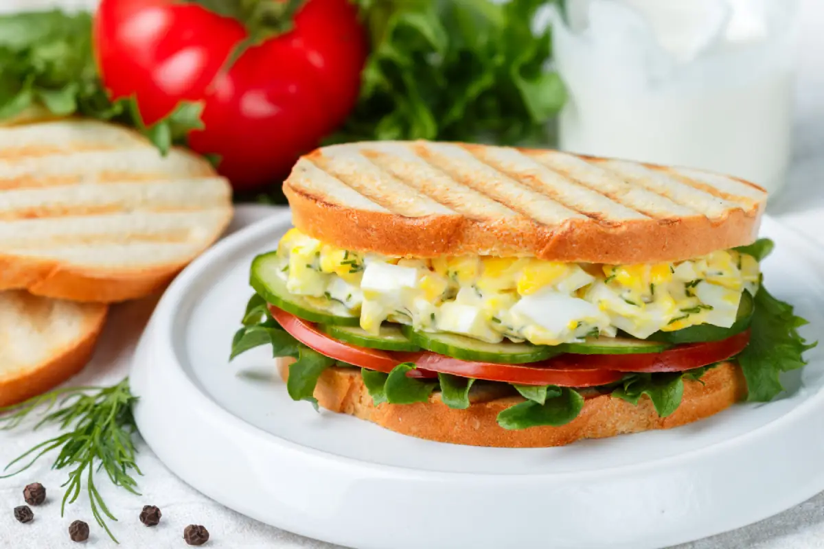 A gourmet egg salad sandwich with fresh lettuce, tomatoes, cucumbers, and dill on grilled bread.