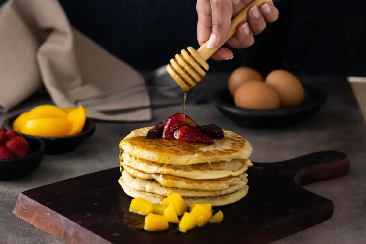 A stack of golden pancakes topped with fresh berries and drizzled with honey, with soft-focus background elements that include whole eggs and sliced mangoes.