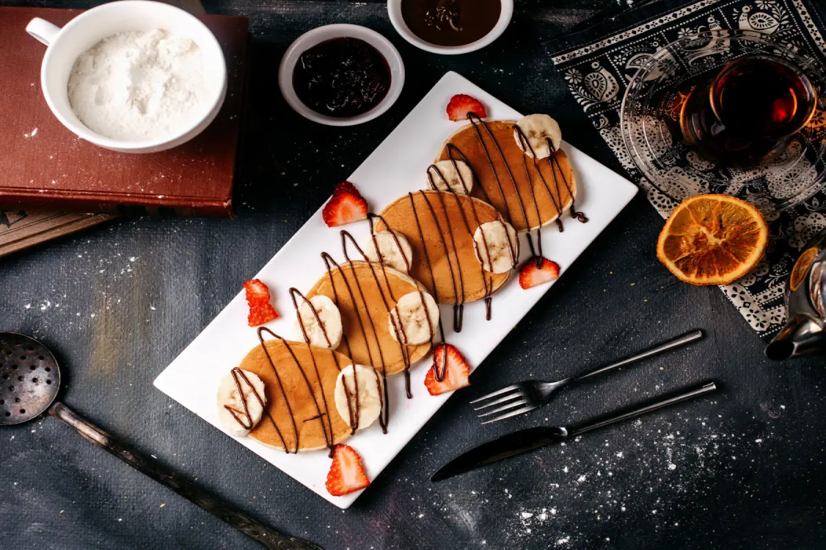 Elegantly plated mini pancakes with chocolate drizzle, accompanied by fresh strawberry slices and banana on a white rectangular dish, with a dark, textured background enhancing the warm tones.