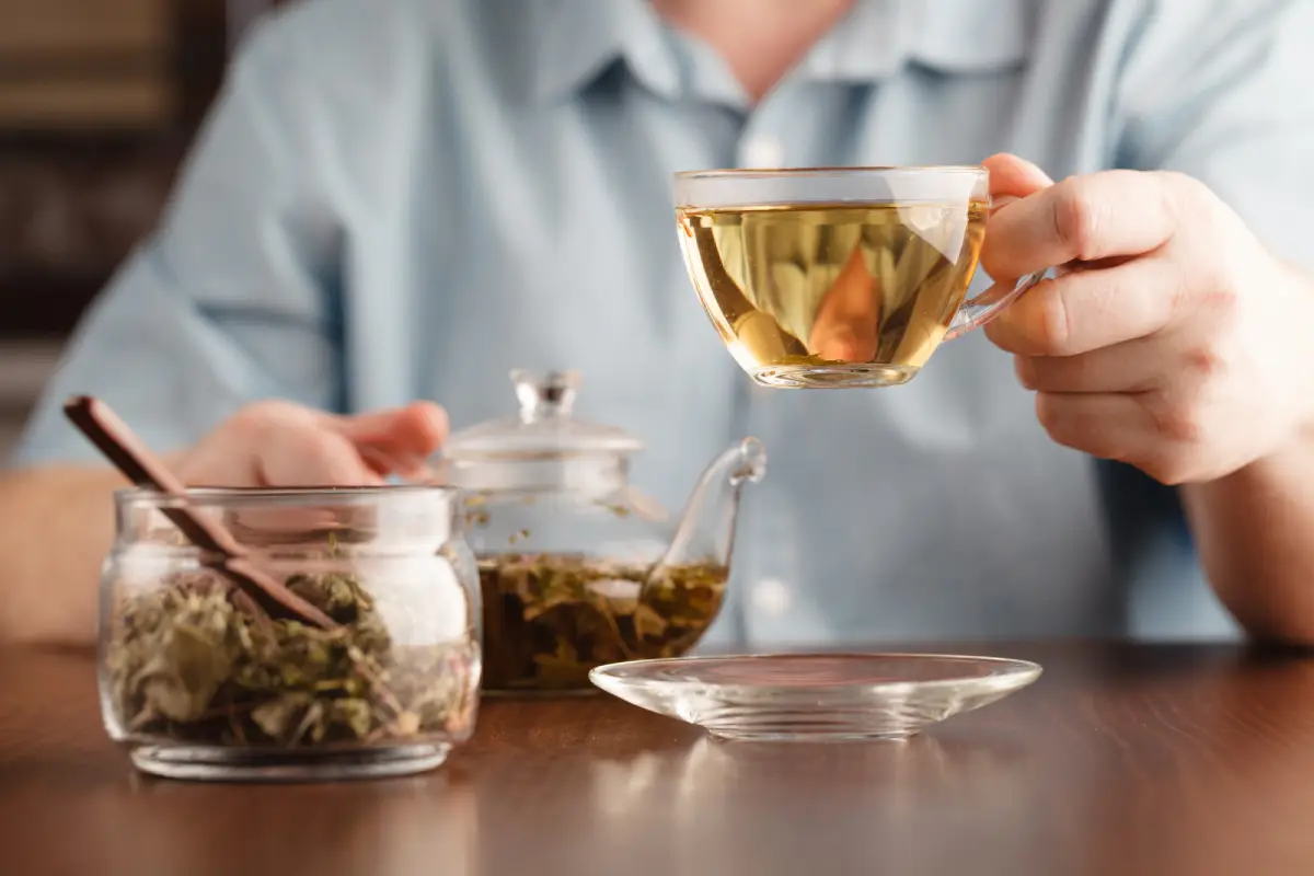 A person holding a clear cup of brewed Earl Grey tea, with a teapot and jar of loose leaves nearby.