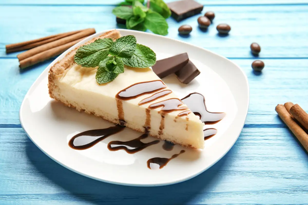A creamy slice of vanilla cheesecake with chocolate sauce and mint on a blue wooden background.