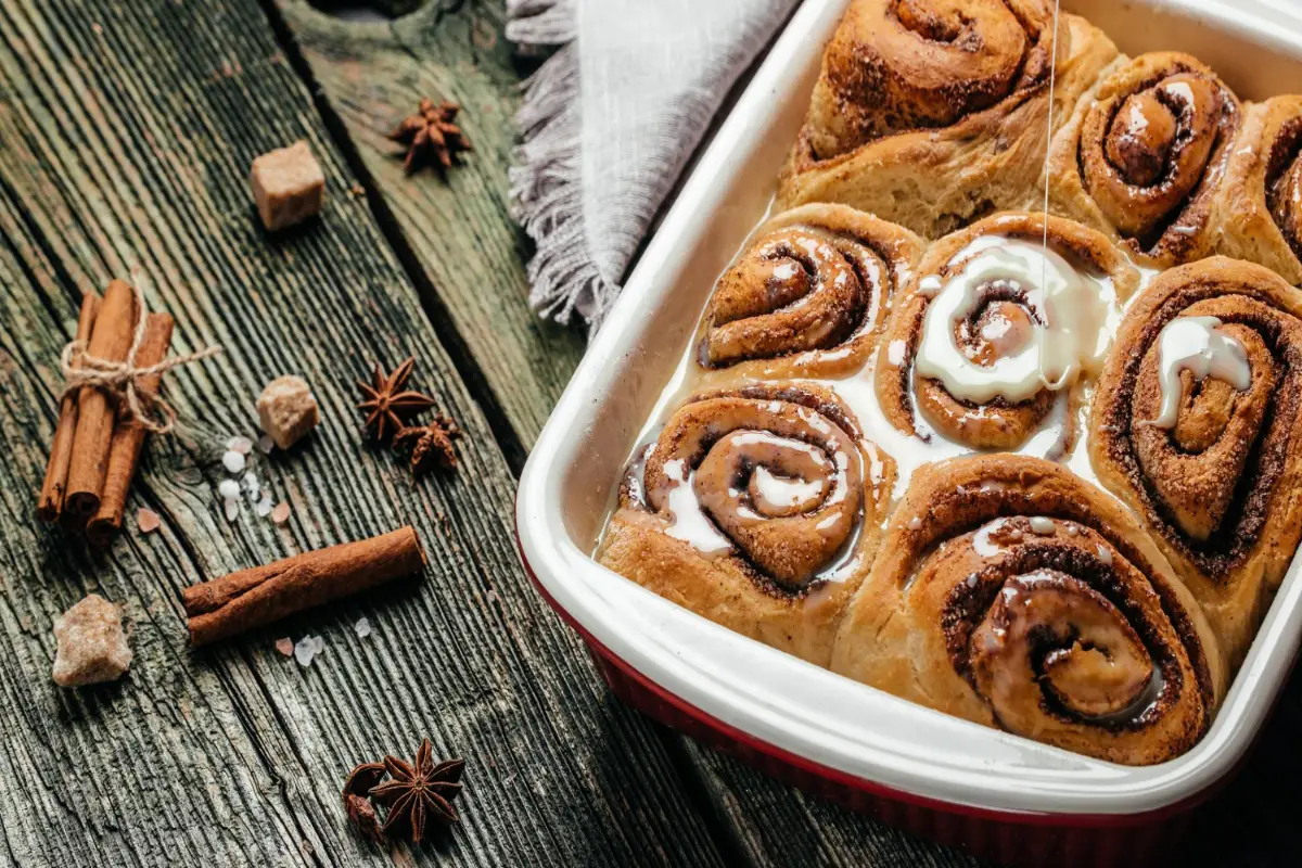 Warm cinnamon rolls in a baking tray with icing and spices on the side.