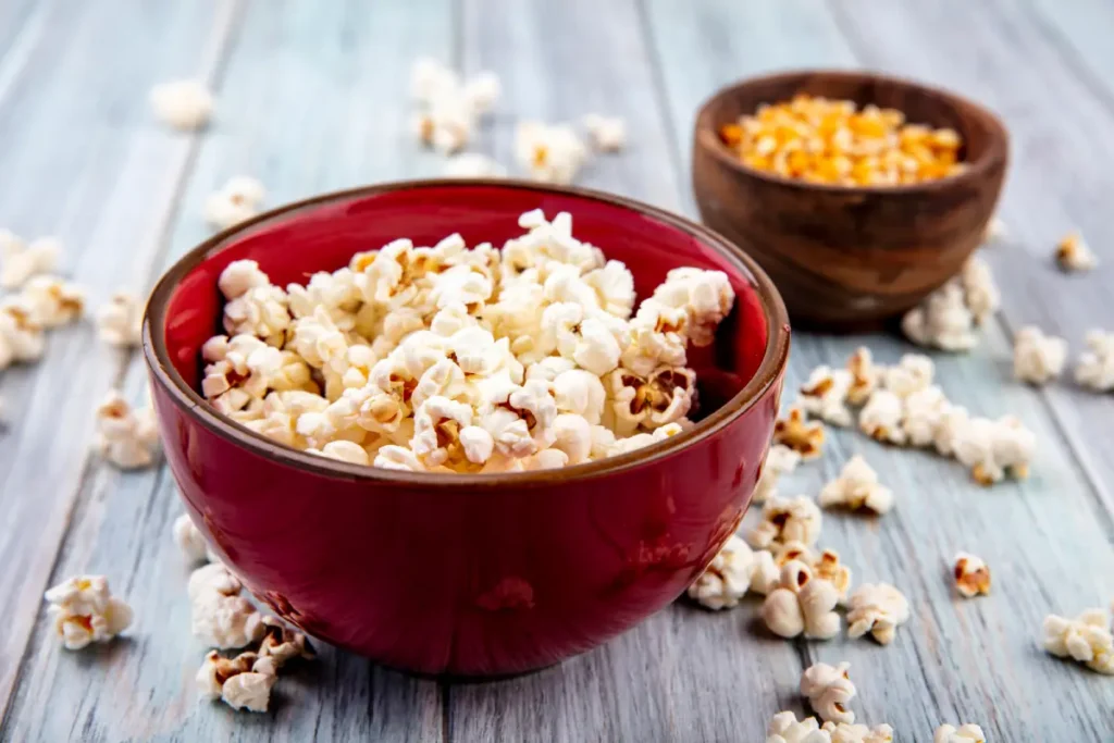 A bowl of freshly popped popcorn next to a bowl of corn kernels on a wooden surface.