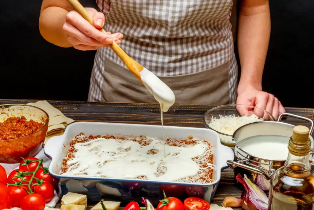 A person adding a creamy béchamel sauce over a lasagna layer, preparing to achieve the optimal lasagna layers with fresh ingredients surrounding the baking dish.