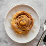 A single cinnamon roll topped with icing on a white plate with utensils on the side.