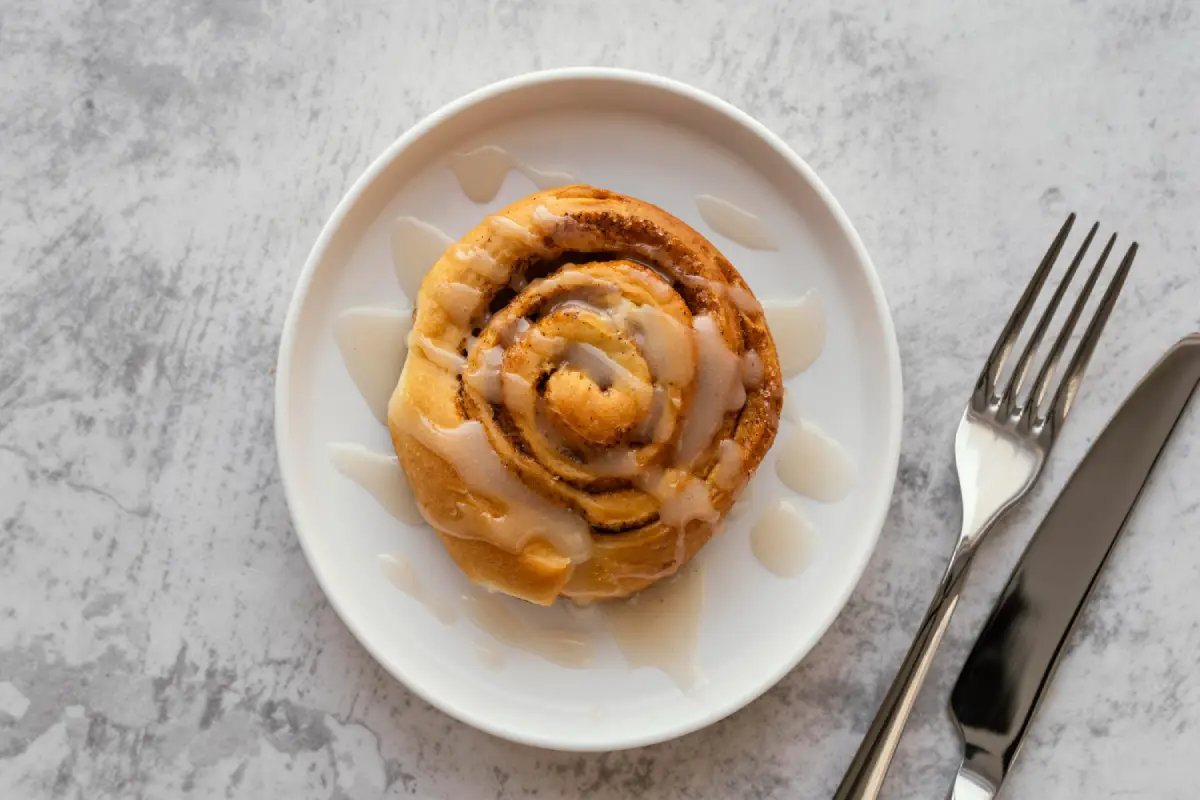 A single cinnamon roll topped with icing on a white plate with utensils on the side.