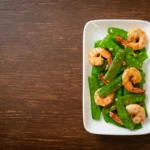 A delectable serving of stir-fried green peas with shrimp on a white plate.